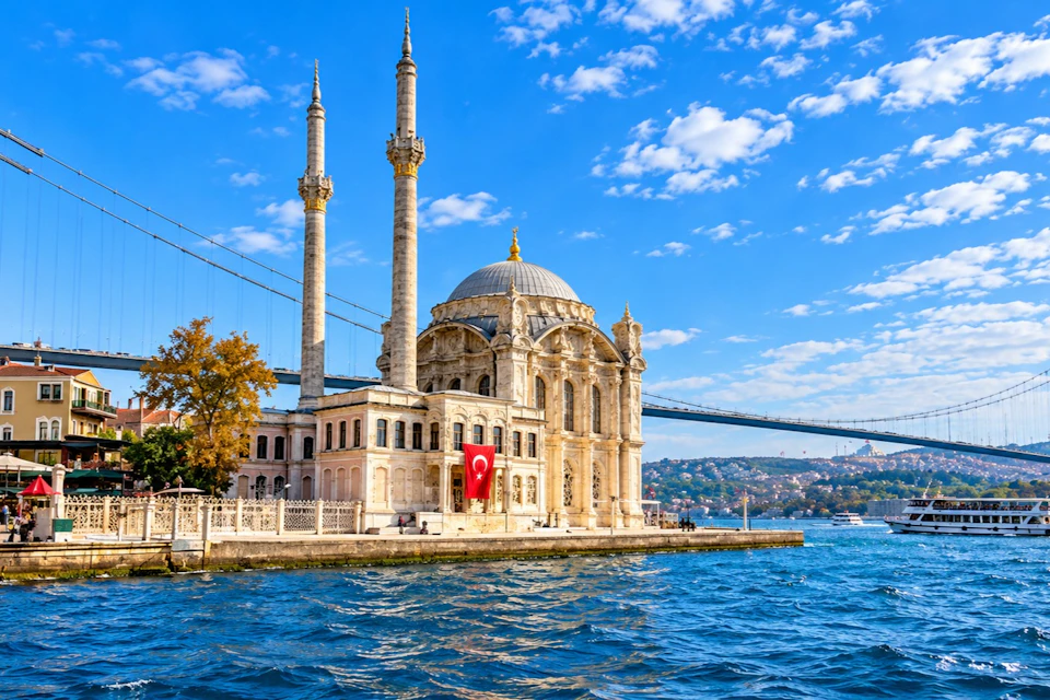 A mosque with two minarets by the water, featuring a Turkish flag, with a bridge and a cityscape in the background.
