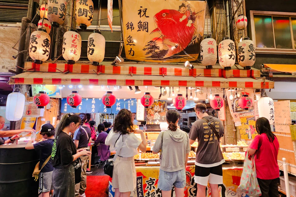 People browsing food stalls decorated with lanterns and signs at a bustling market.