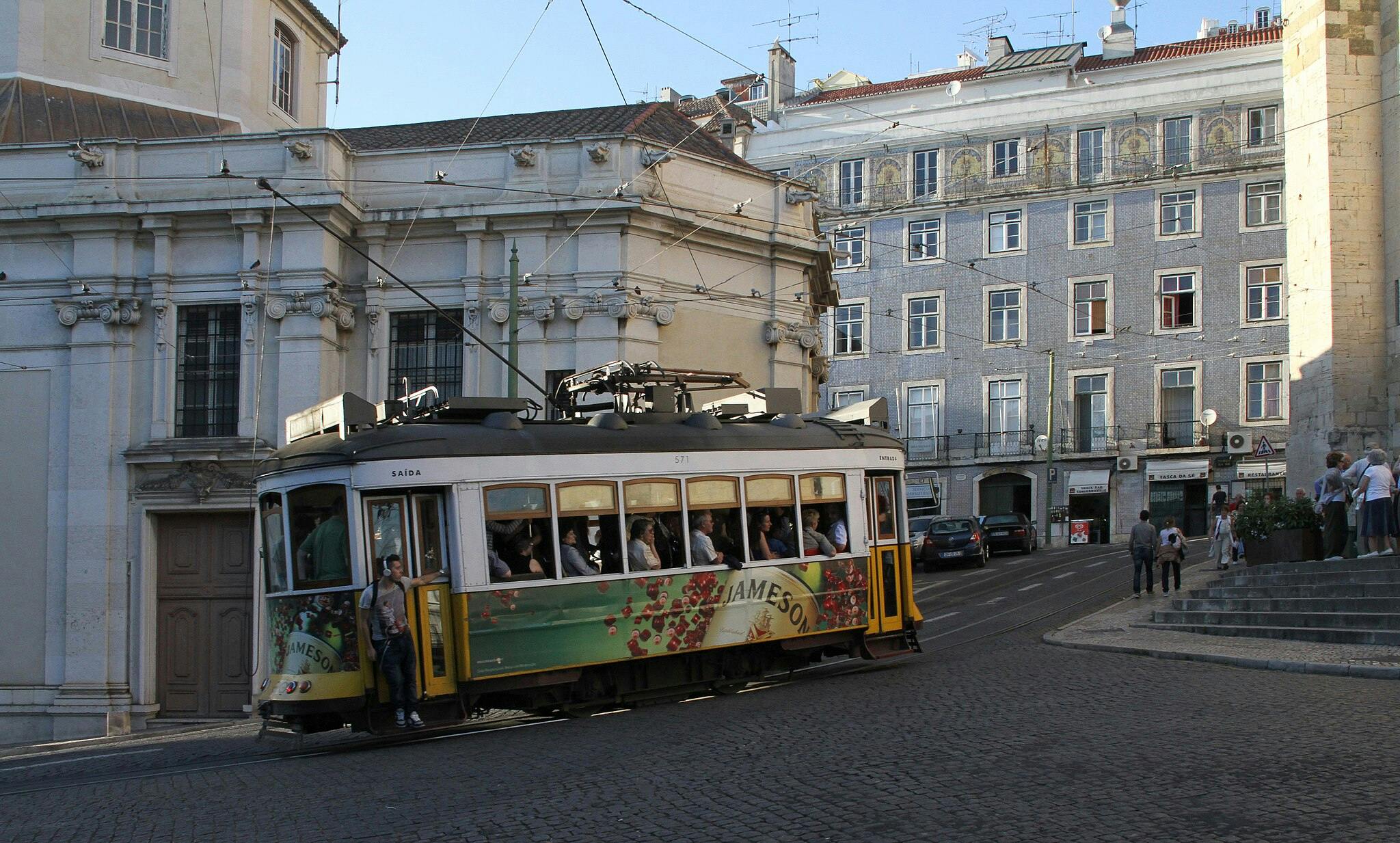 A vintage tram with passengers travels up a sloped street between historic buildings. People walk on the sidewalk nearby.