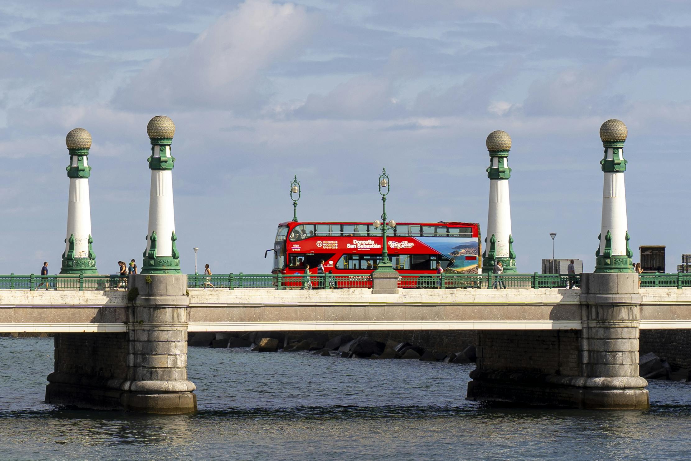 People sitting on a double-decker bus, viewing a scenic beach with a hill in the background under a clear blue sky.