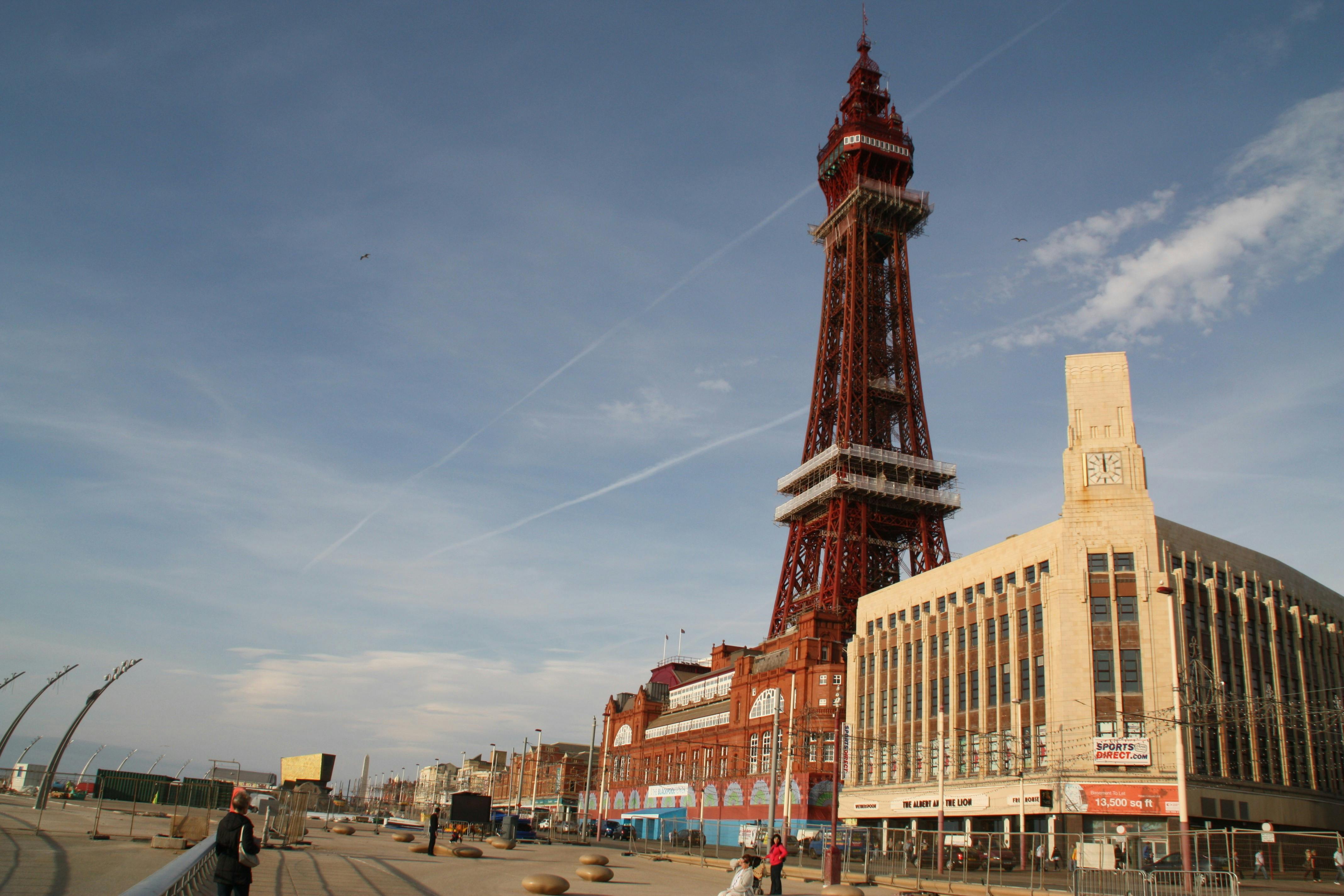 Tall red tower with a lattice structure, surrounded by buildings, under a clear sky; few people walk nearby.