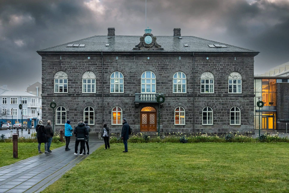 A group of people gather outside a large, two-story stone building with arched windows under a cloudy sky.