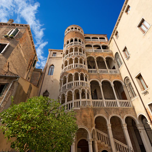 Hög italiensk byggnad med många valv och balkonger mot en blå himmel, omgiven av andra äldre byggnader och ett träd.