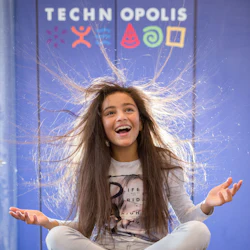 En tjej med håret stående i änden sitter med benen i kors och ler mot Technopolis.