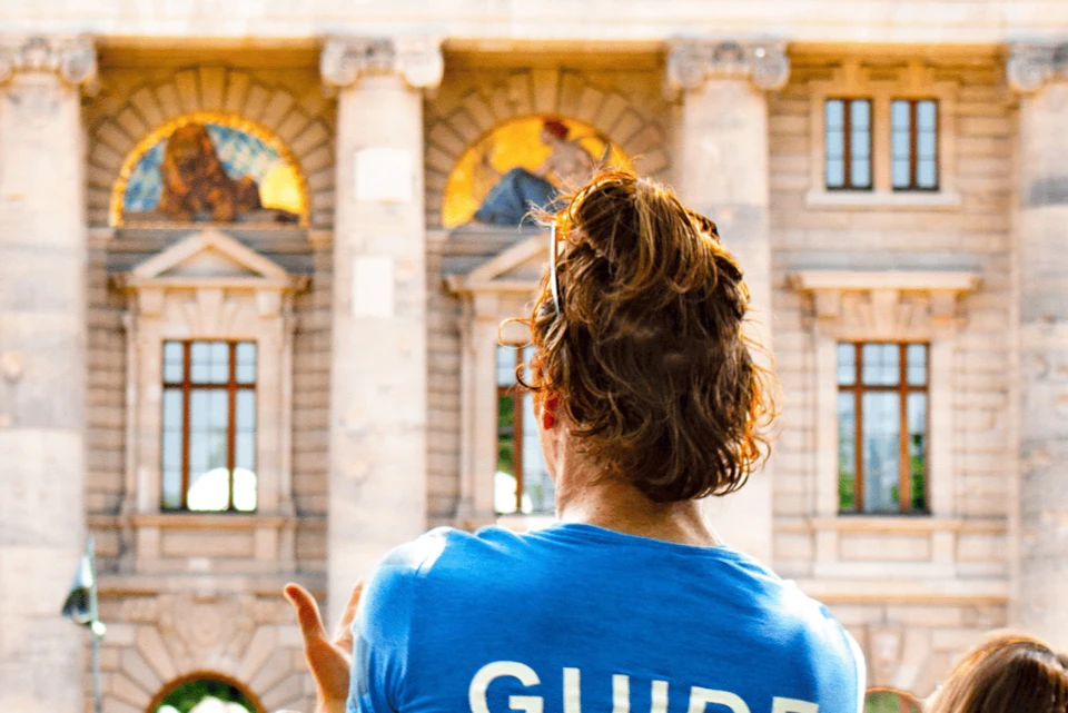 Person with curly hair in a blue shirt labeled "GUIDE," gesturing in front of a historic building with large columns and mosaics.