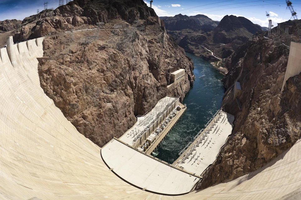 A view of a large concrete dam surrounded by rocky cliffs, overlooking a river with a power plant at the base.