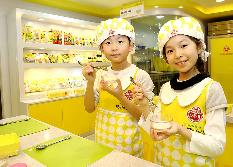 Two children in matching yellow aprons and chef hats holding small bowls and spoons in a brightly lit kitchen.