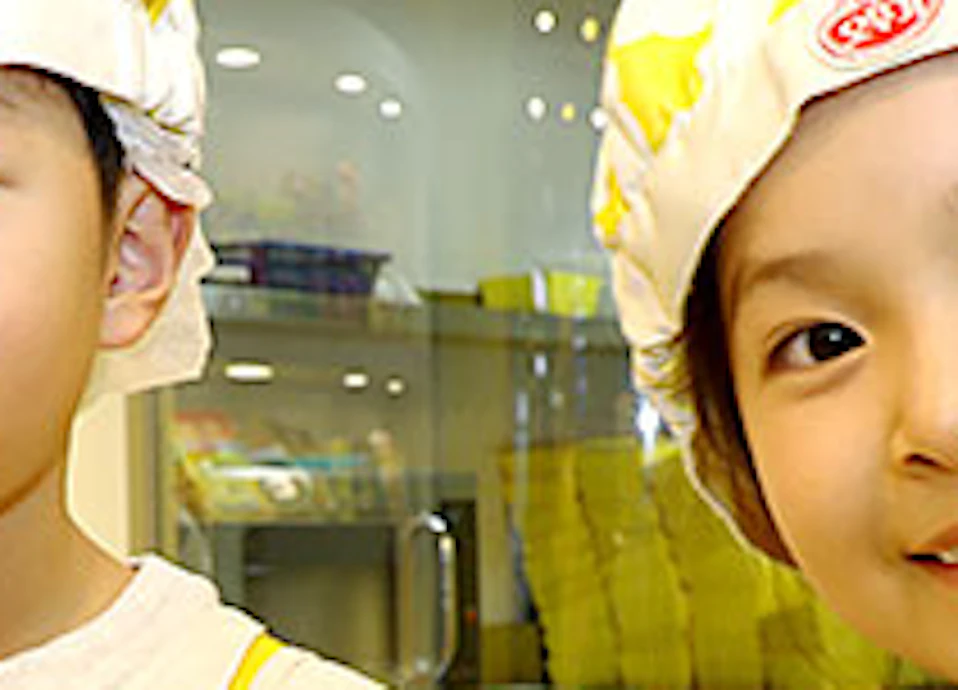 Two children in matching yellow aprons and chef hats holding small bowls and spoons in a brightly lit kitchen.