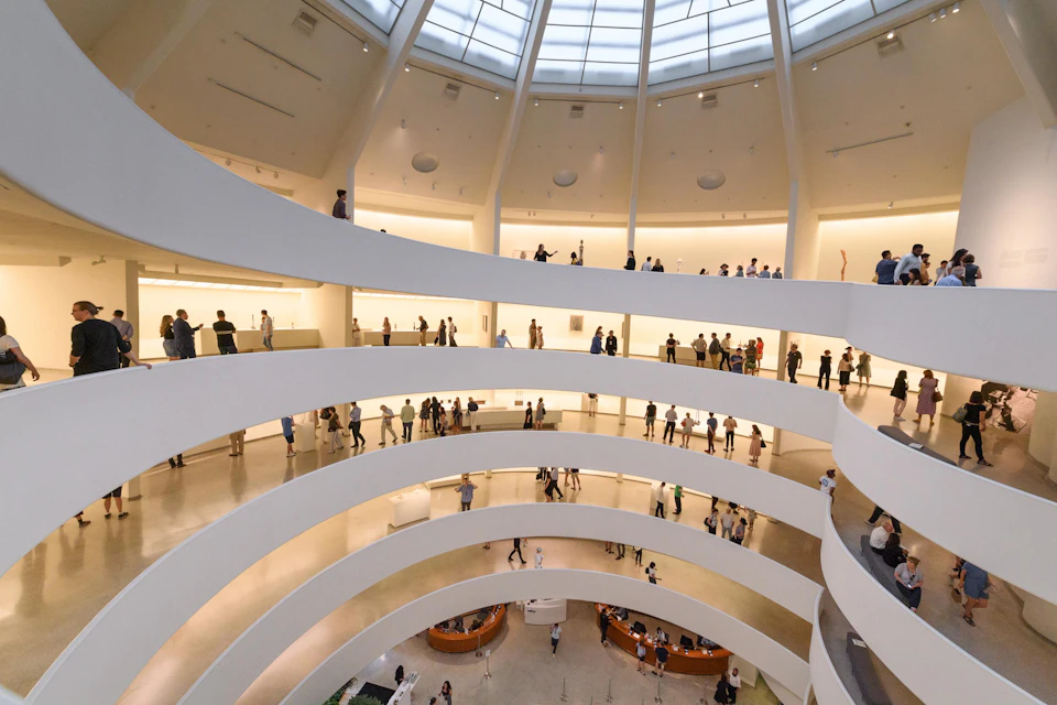 Interior of a multi-level museum with numerous visitors walking along circular ramps and viewing exhibits under a large skylight.