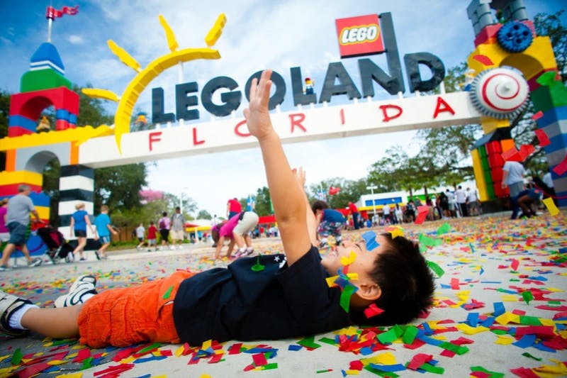 Ett barn ligger på ett golv täckt med färgglada konfetti under Legoland Floridas entréskylt. Familjer finns i bakgrunden.