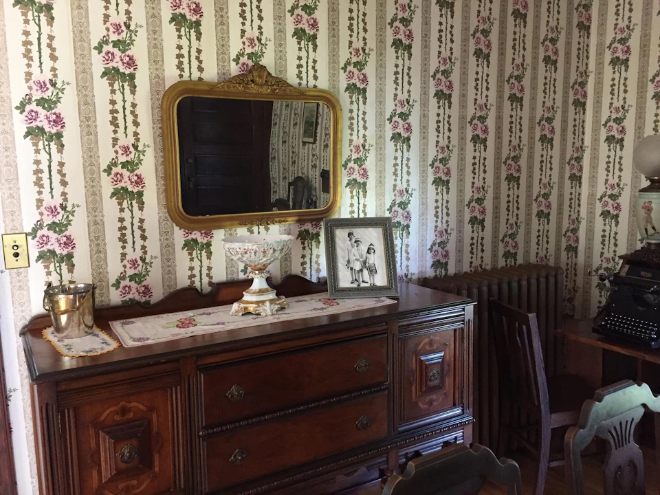 Room with floral wallpaper, a wooden dresser with a doily, a framed black-and-white photo, a decorative bowl, and a large mirror.