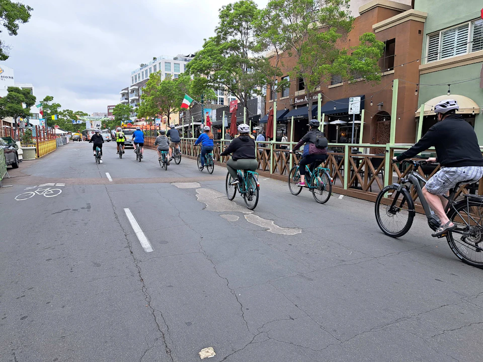 A group of cyclists rides down an urban street with restaurants and trees lining the sidewalk.
