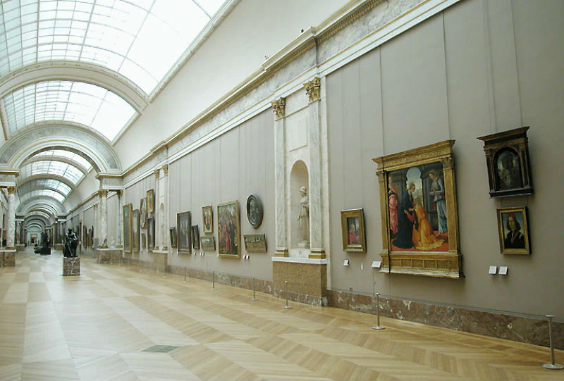 A spacious gallery with paintings and sculptures displayed on both sides of a long, well-lit hallway with a high arched ceiling.
