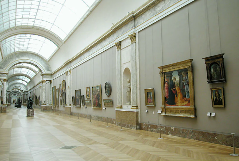A spacious gallery with paintings and sculptures displayed on both sides of a long, well-lit hallway with a high arched ceiling.