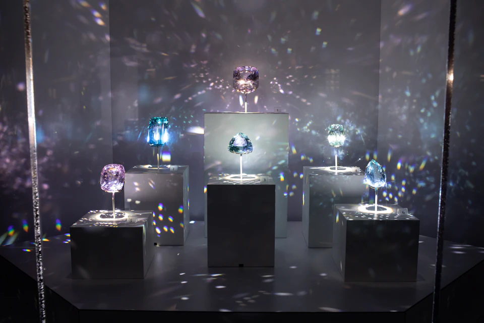 Six illuminated colored crystals are displayed on white pedestals in a dim room, with light reflecting off them creating a sparkling effect.