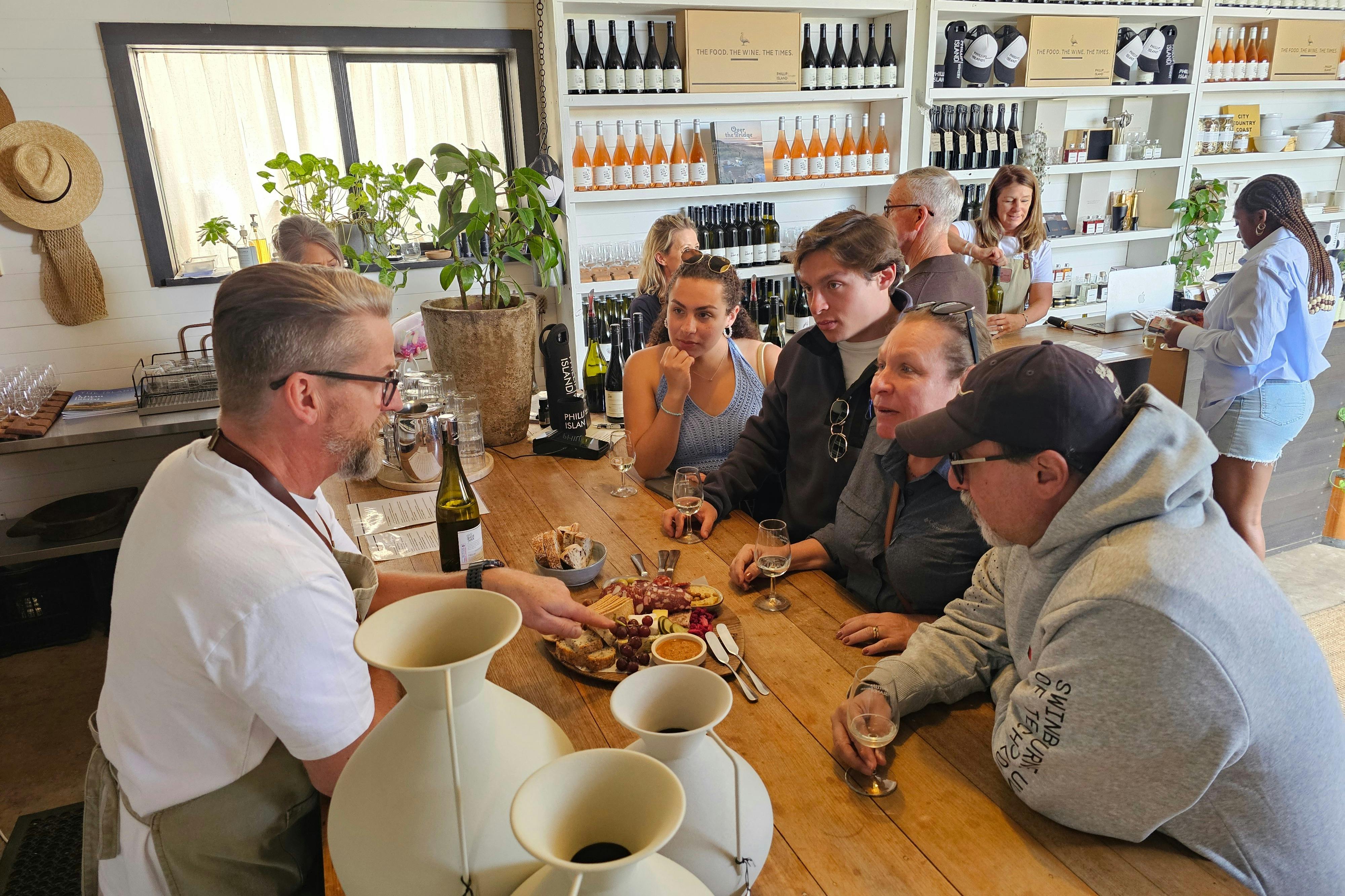 A man explains a charcuterie board to four people at a wine tasting bar with shelves of wine bottles in the background.