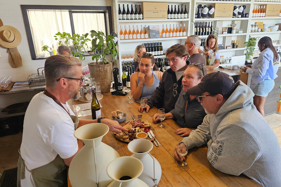 A man explains a charcuterie board to four people at a wine tasting bar with shelves of wine bottles in the background.