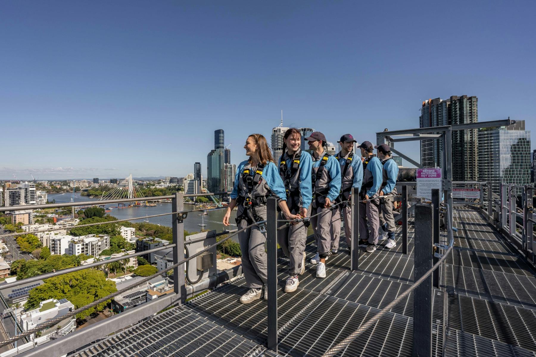 一群人穿着马具在城市景观上方的金属人行道上行走，那里有高楼大厦和河流。