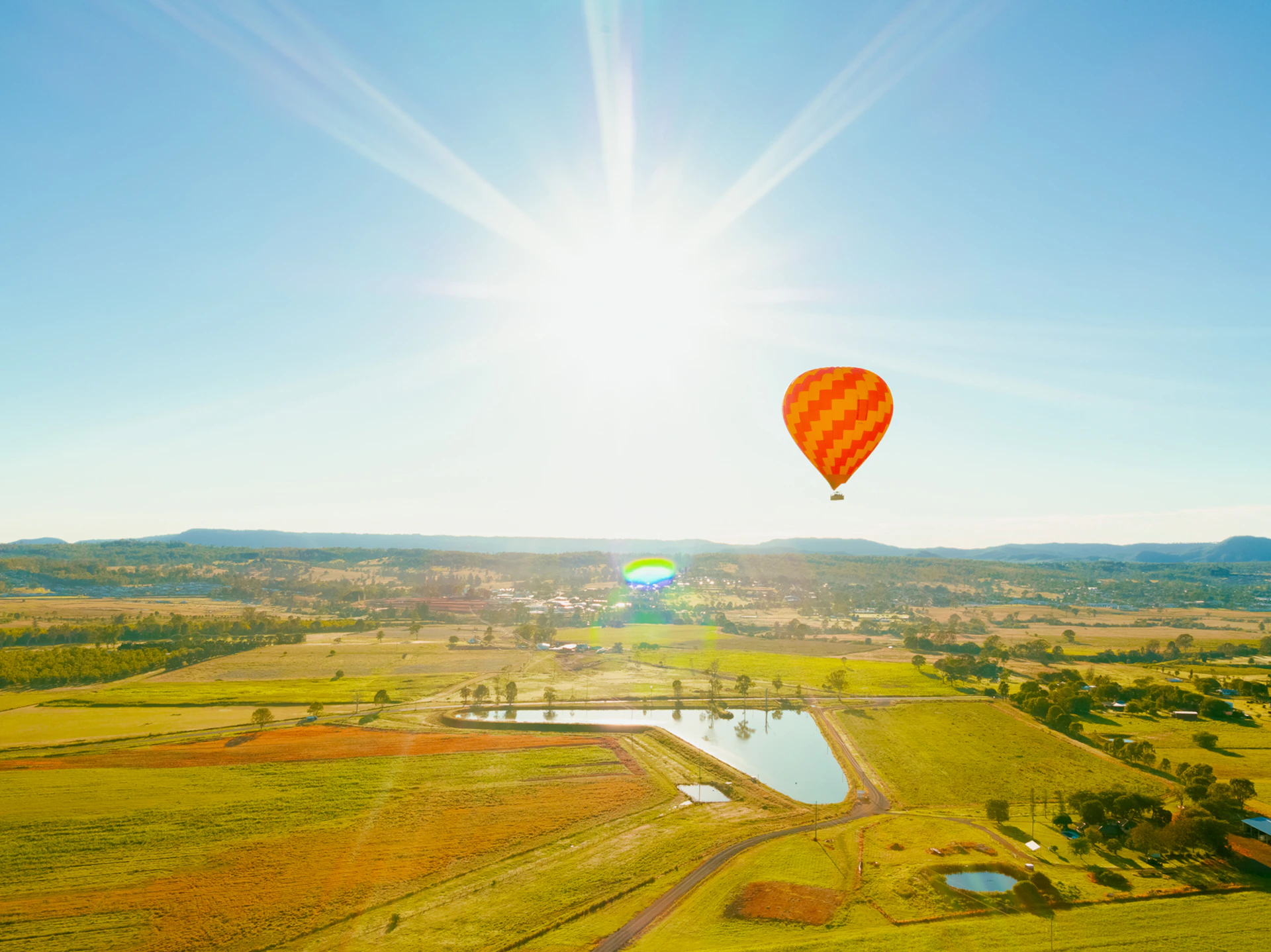 Three hot air balloons, one large yellow with "Austra" text, and two smaller ones, float above a scenic landscape with hills and trees.