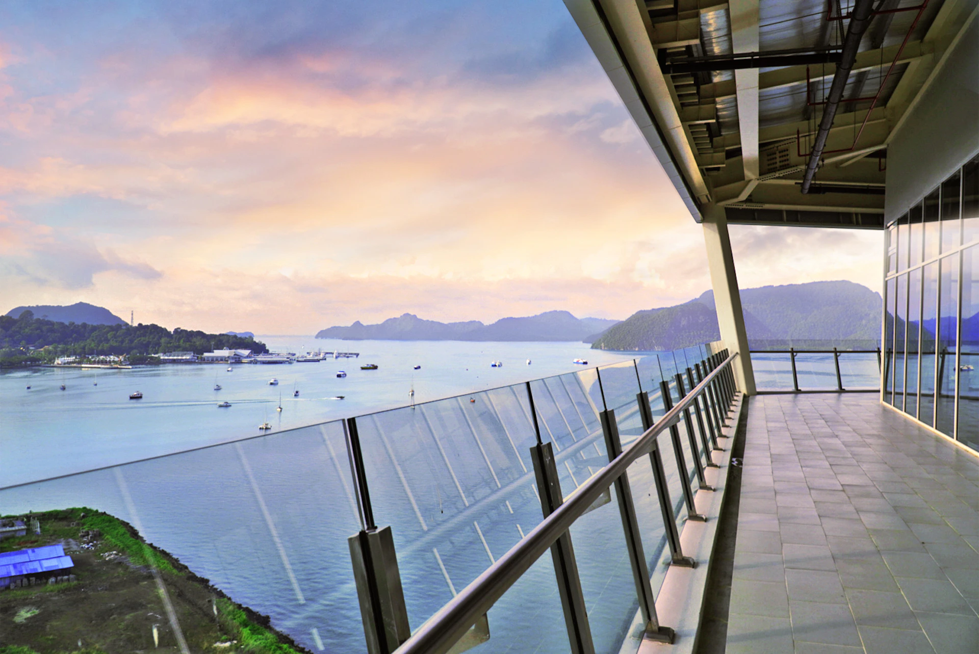 A balcony with a glass railing overlooking a calm sea with boats and distant hills under a colorful sky.