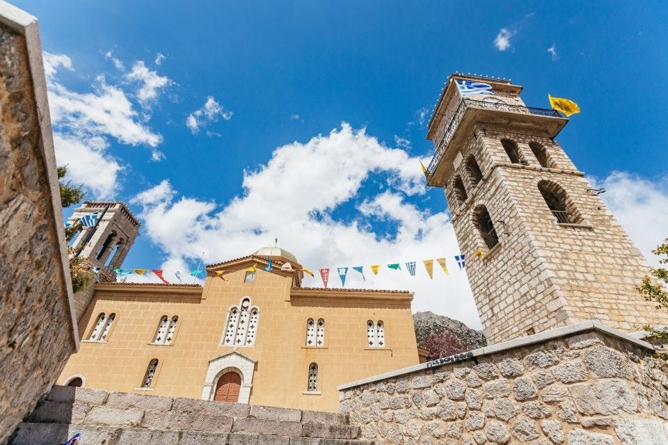 아치형 창문과 종탑이 있는 노란색 석조 건물로, 구름과 화려한 깃발이 있는 푸른 하늘을 배경으로 합니다.