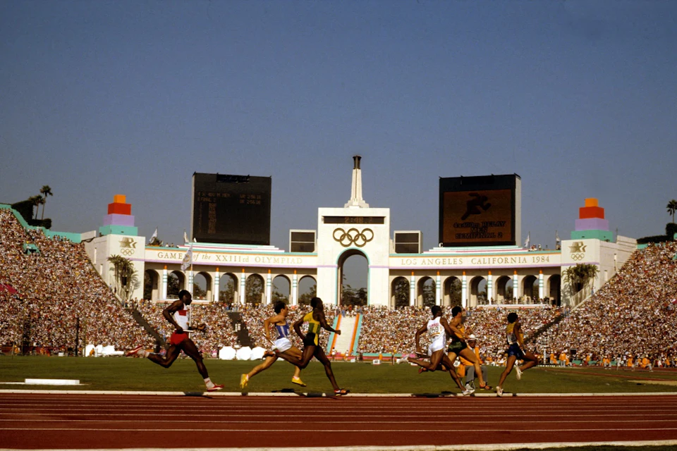 Sprinters competing in a race at the 1984 Los Angeles Olympics, with a packed stadium and Olympic rings visible in the background.