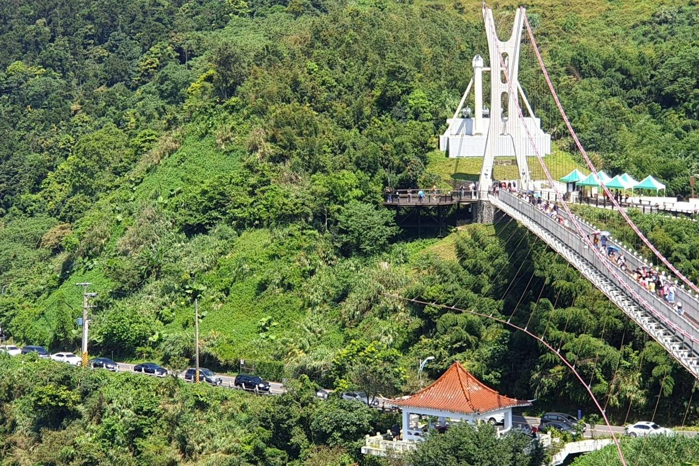 A suspension bridge spans a green, forested valley, with a red-roofed structure and parked cars below.