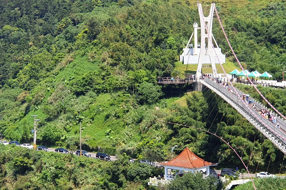 A suspension bridge over a lush green valley with a red-roofed pavilion and cars on a nearby road.