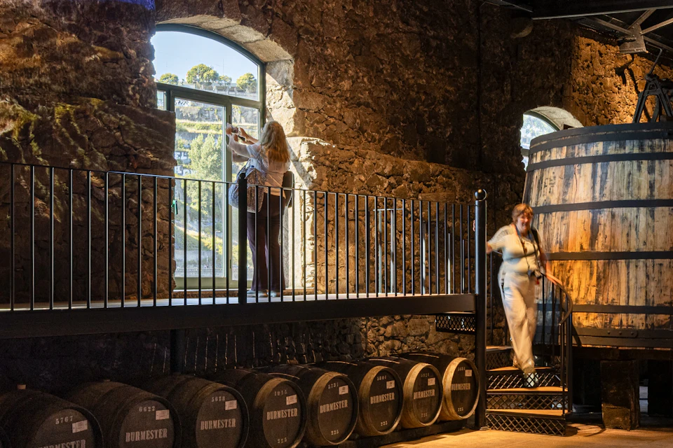 A stone-walled room with wine barrels labelled ‘Burmester’, a person at a window, and another person coming down the stairs.