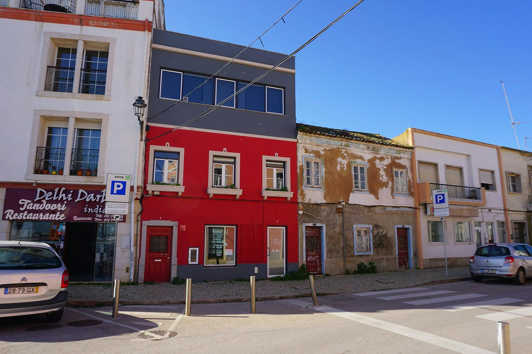 Una escena al carrer amb edificis de colors i condicions mixtes, cotxes aparcats a prop de cartells i un restaurant indi a l'esquerra.