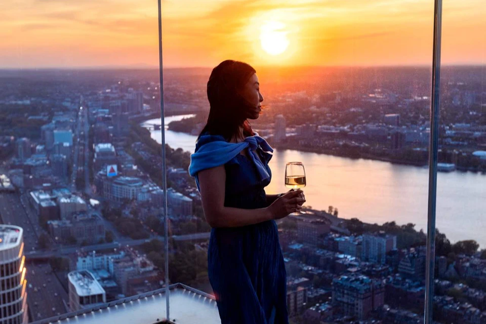 A woman in a blue dress holds a glass of wine while looking out at a cityscape and river during sunset from a tall building.