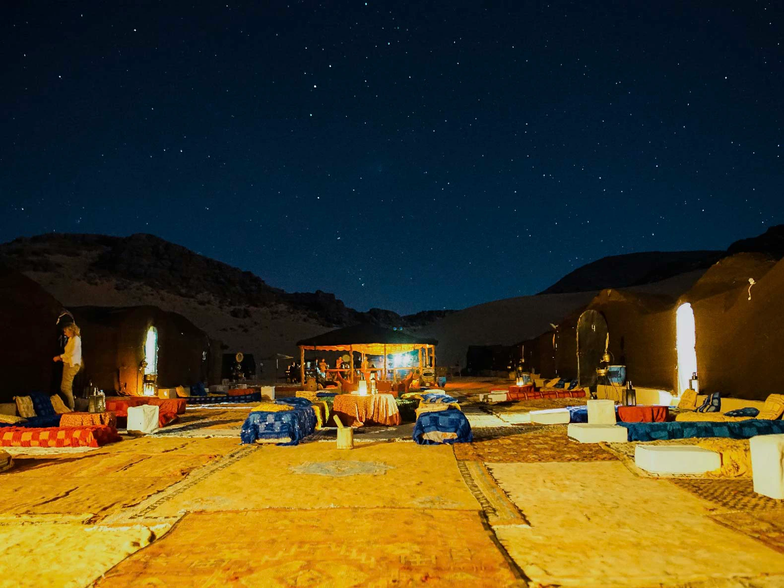 Outdoor seating setup in a desert under a starry night sky. Cushions and low tables are arranged around a central lit area.
