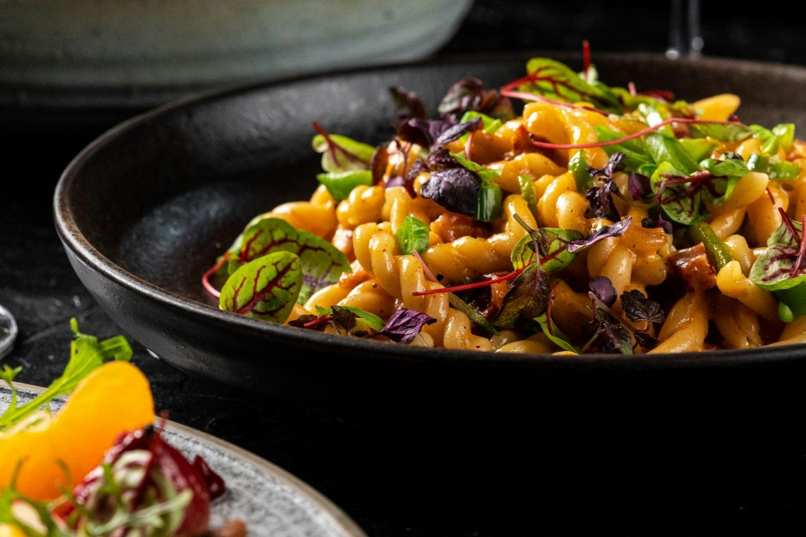 Close-up of a black bowl containing pasta garnished with fresh greens and herbs.