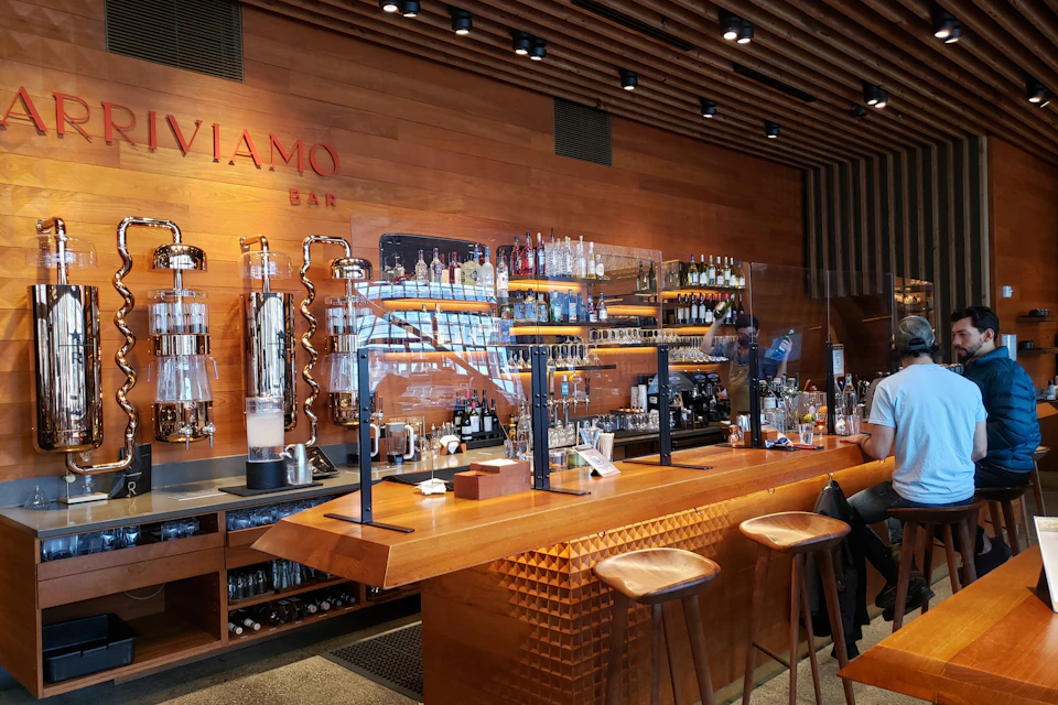 Wooden bar with high stools, a variety of liquor bottles on shelves, and coffee equipment behind a plexiglass partition.