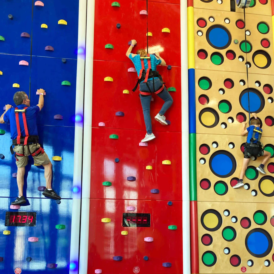 Three people in harnesses climbing brightly colored indoor rock walls; one blue, one red, one yellow with circles.