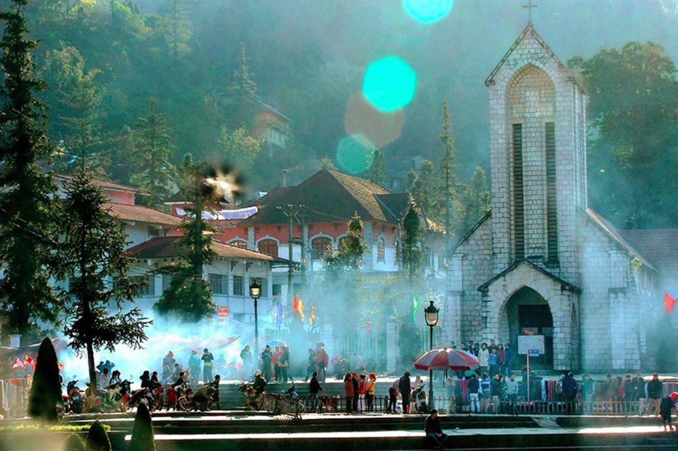 A busy town square with people walking and cycling, featuring a stone church, smoke rising, and surrounding trees and buildings.
