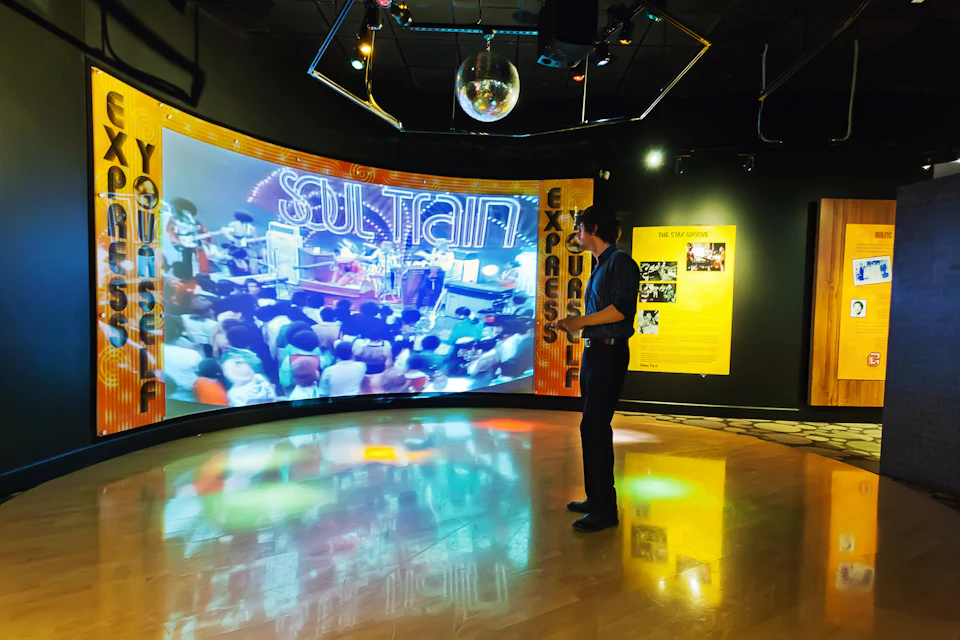 A person stands in an exhibit room watching a large screen displaying a "Soul Train" video under a disco ball.