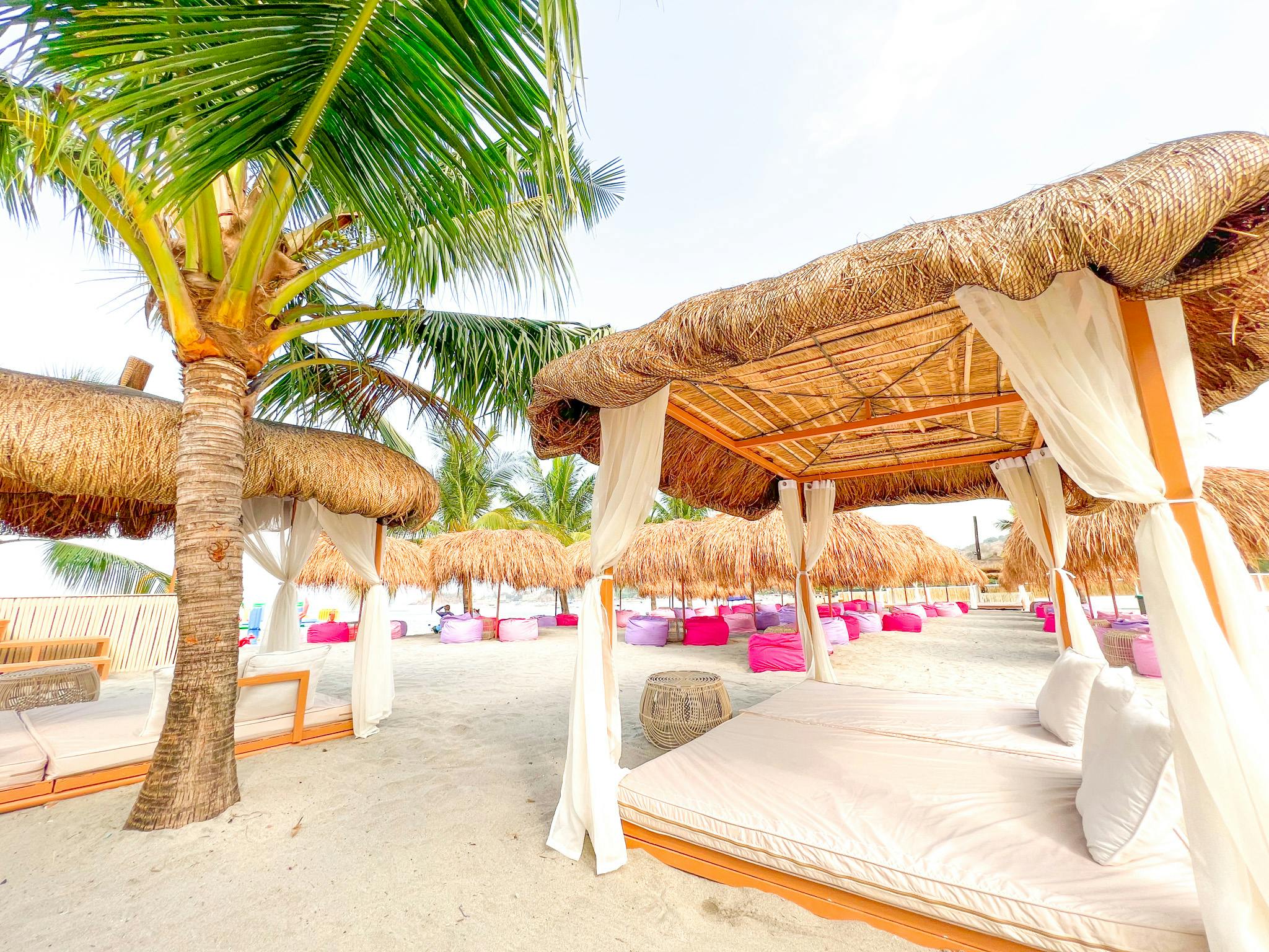 A tropical beach scene with palm trees, thatched cabanas, cushioned seating, and soft sand under a bright sky.
