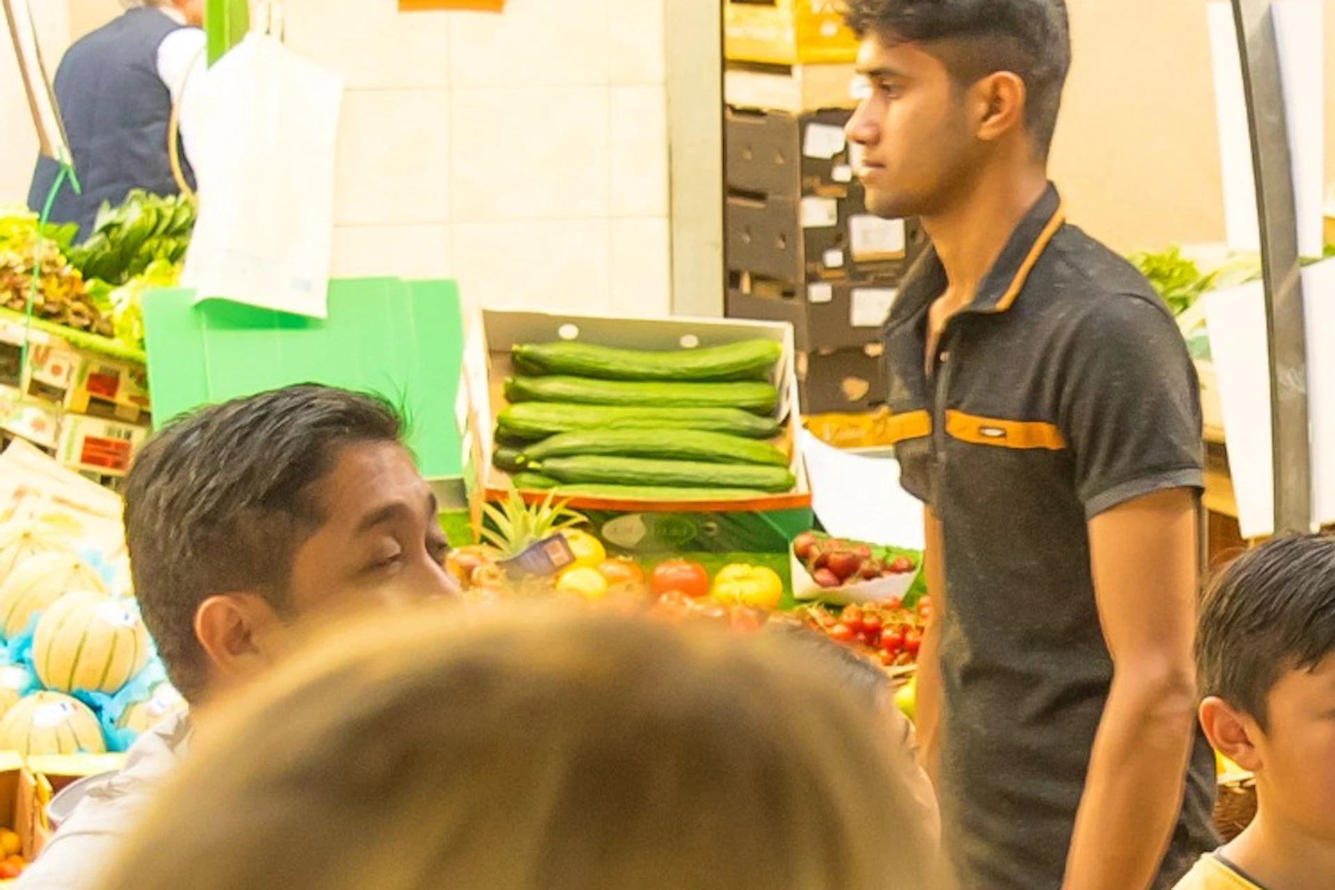A group of people gathered around a man speaking in a market with fresh produce and stalls visible in the background.