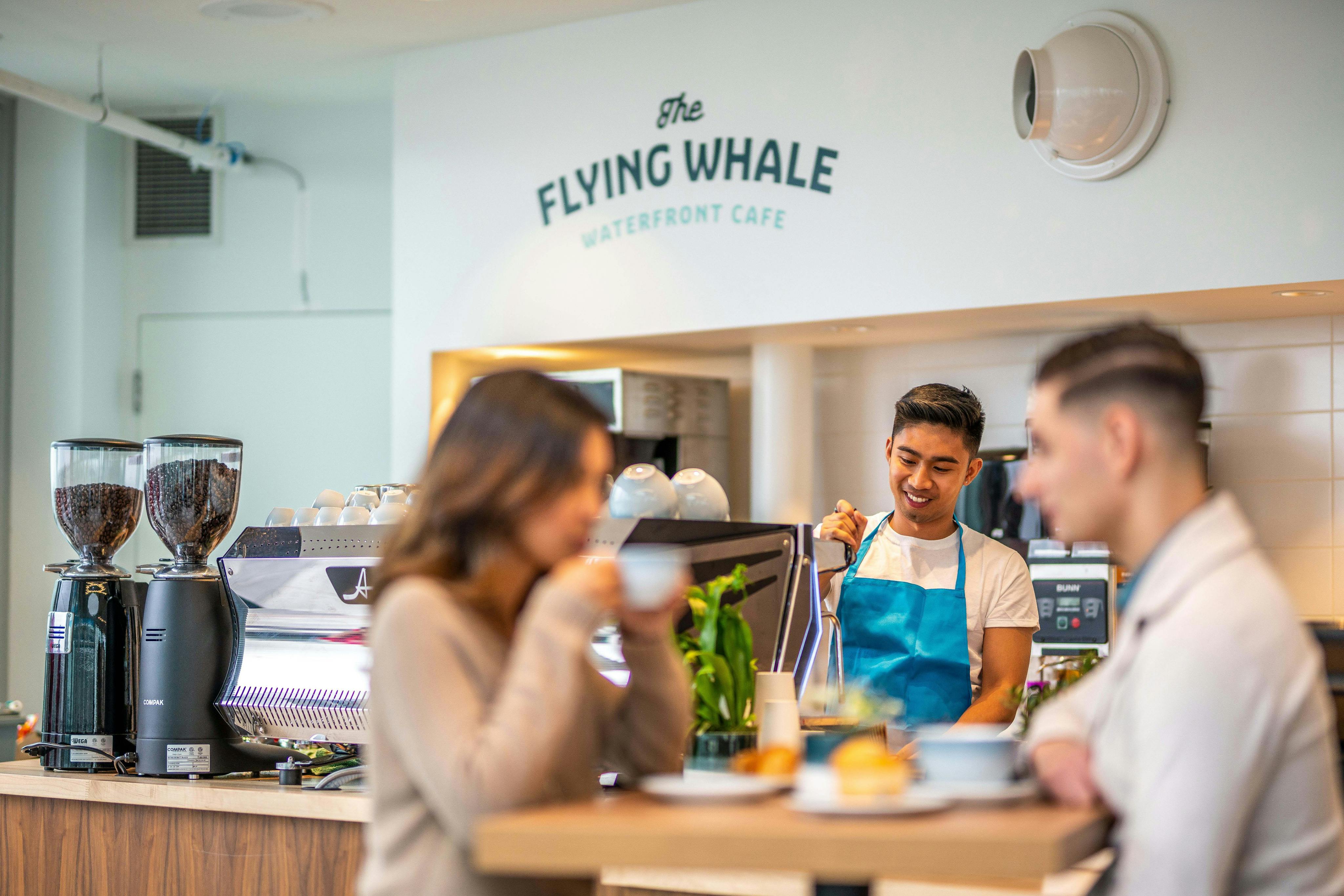 A barista in a blue apron serves drinks at The Flying Whale Café, with two customers seated in the foreground.