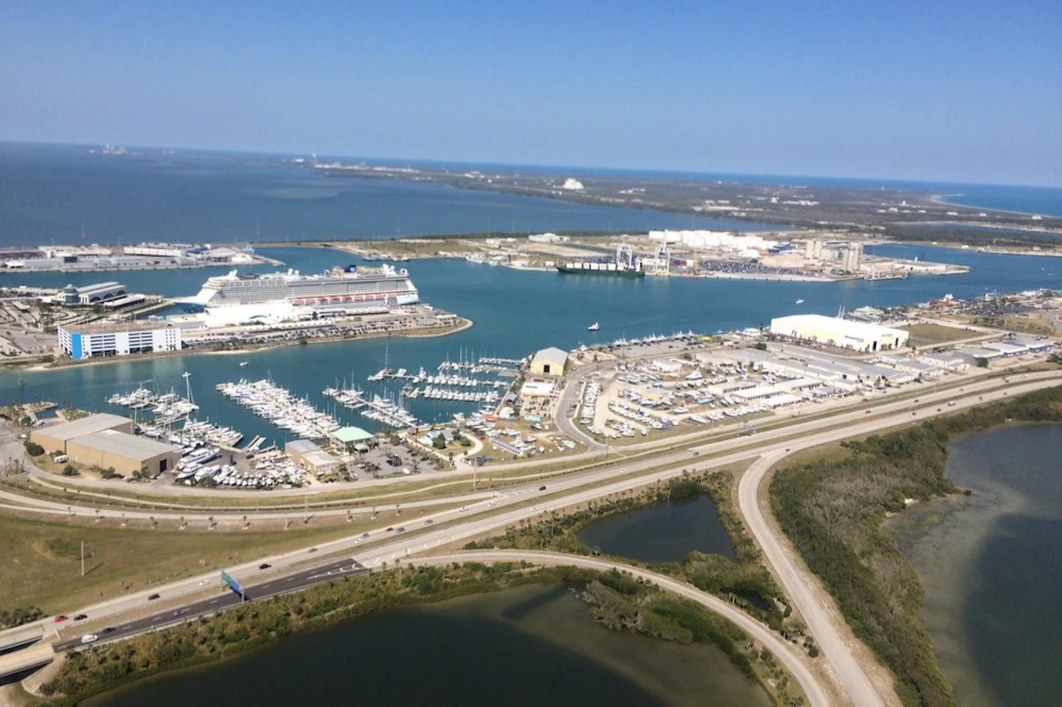 Aerial view of a marina with numerous boats, a cruise ship docked, surrounding buildings, roads, and water in the background.