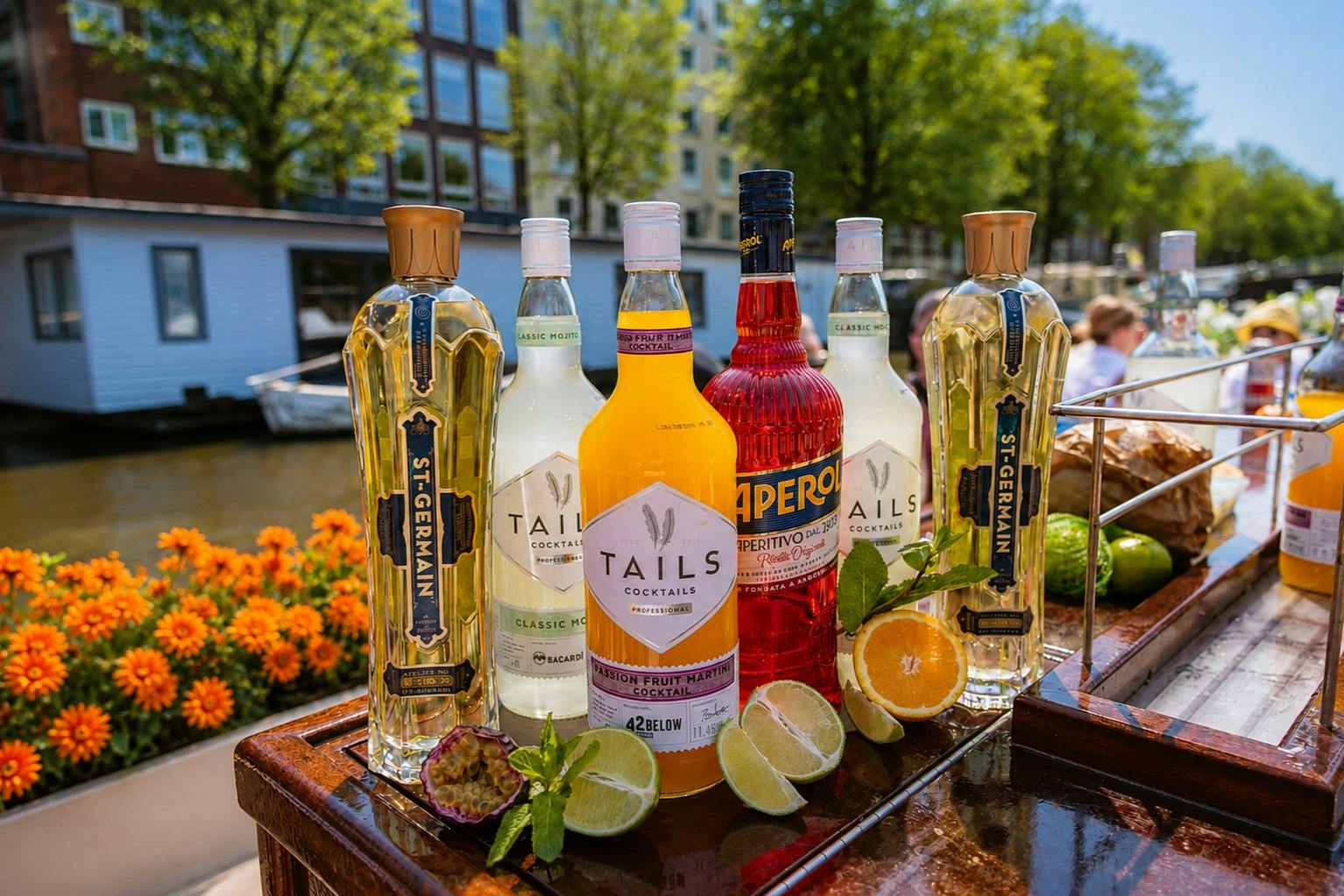 Bottles of various liquors and cocktail mixers displayed on a wooden surface with fruits, set against a canal and greenery.