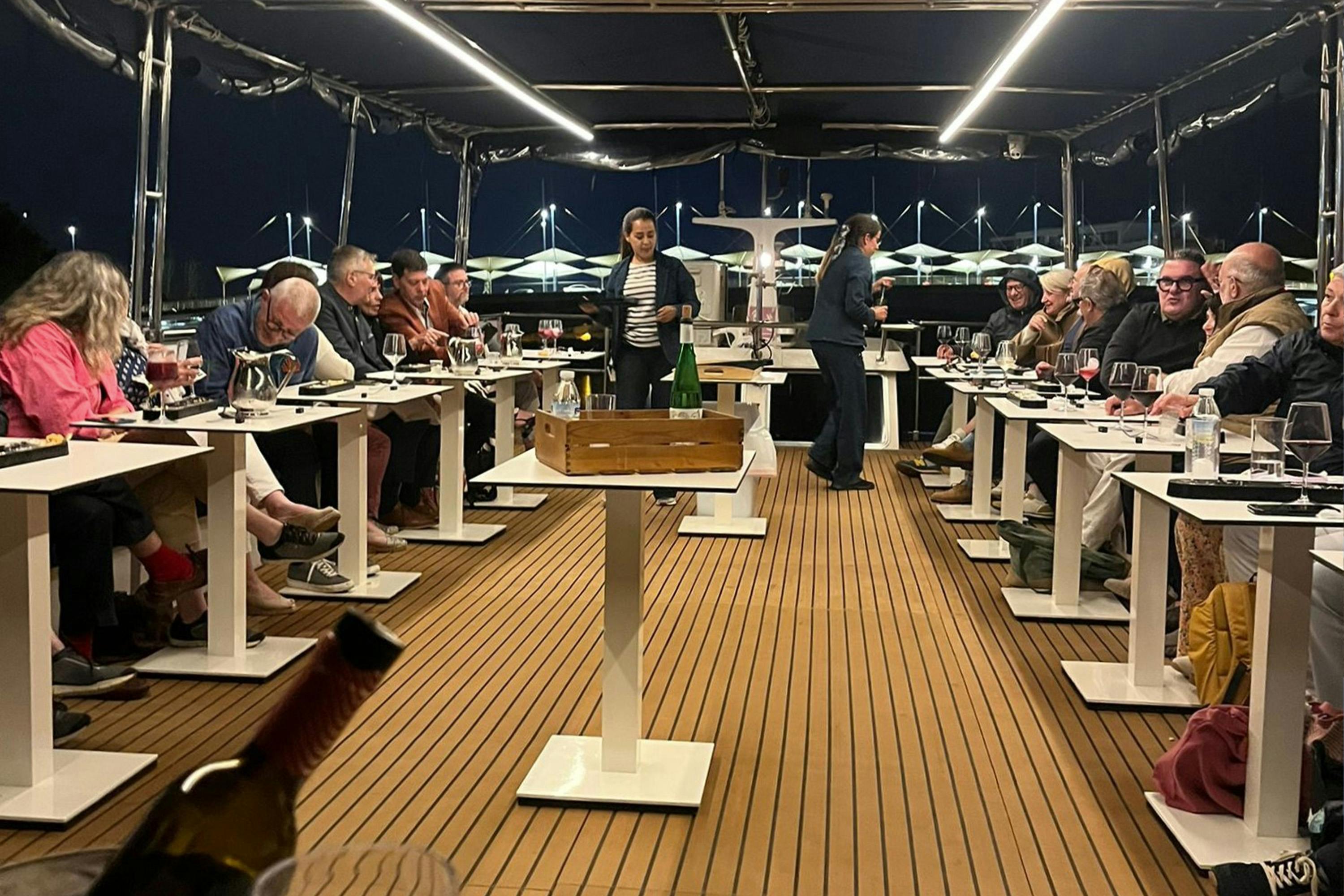 Ludzie siedzący nocą przy stołach na łodzi, zaangażowani w degustację wina, z dwoma kelnerami pośrodku.