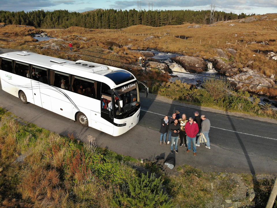 A group of people standing near a white tour bus parked on a narrow road beside a scenic river with falls and grassy hills.