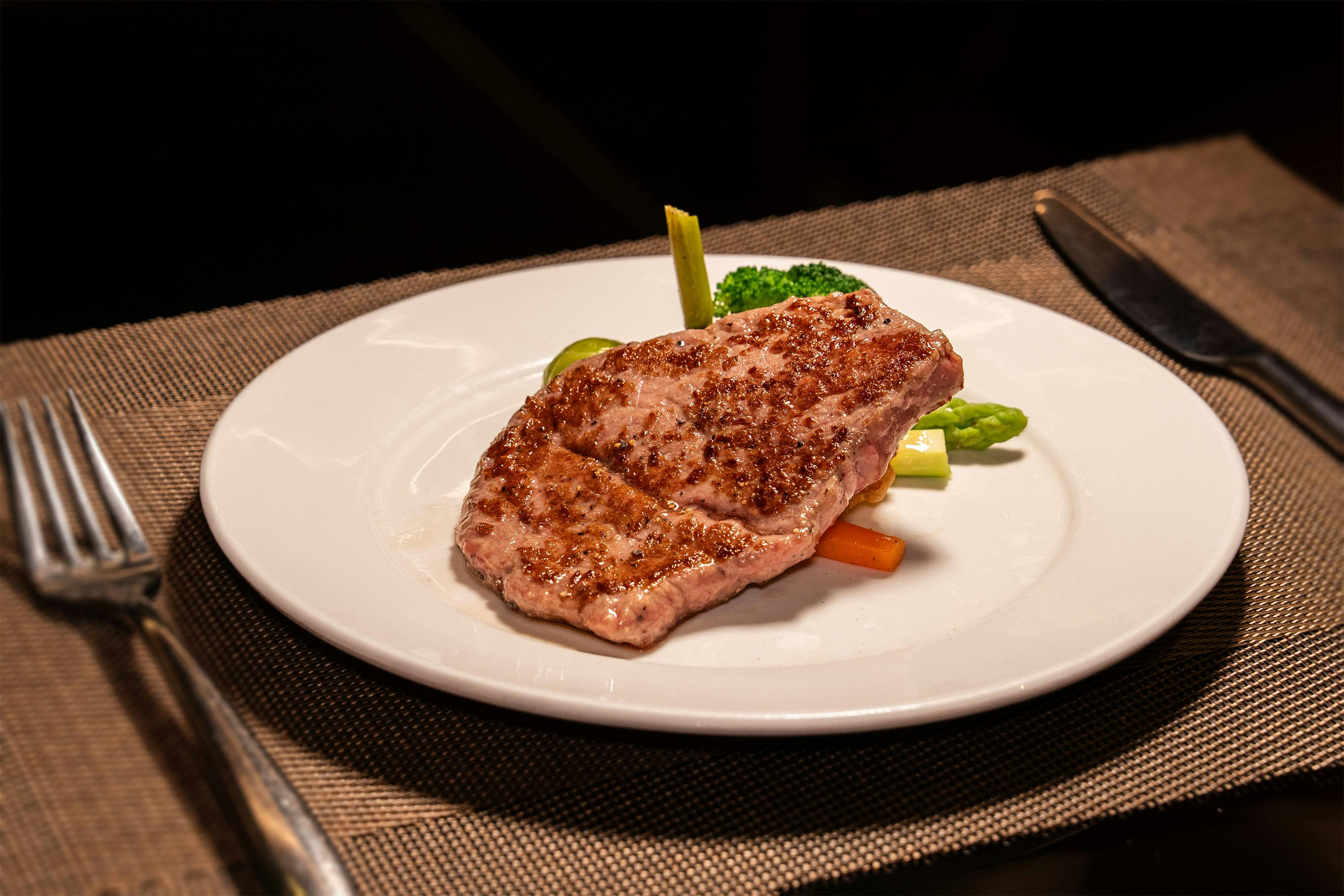 A grilled steak served on a white plate with assorted vegetables, including broccoli and asparagus, set on a placemat.