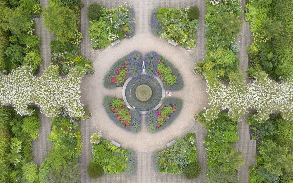Aerial view of a circular garden fountain surrounded by pathways, benches, and lush greenery with symmetrical flower beds.