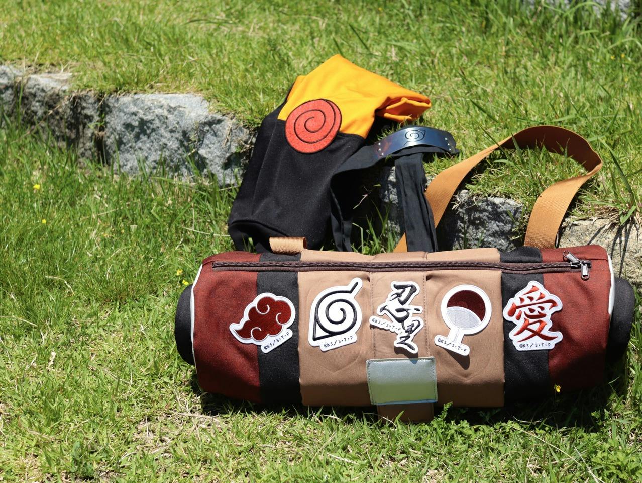 A colorful bag and accessories with anime designs lie on grass near stone steps, featuring various symbols and logos.