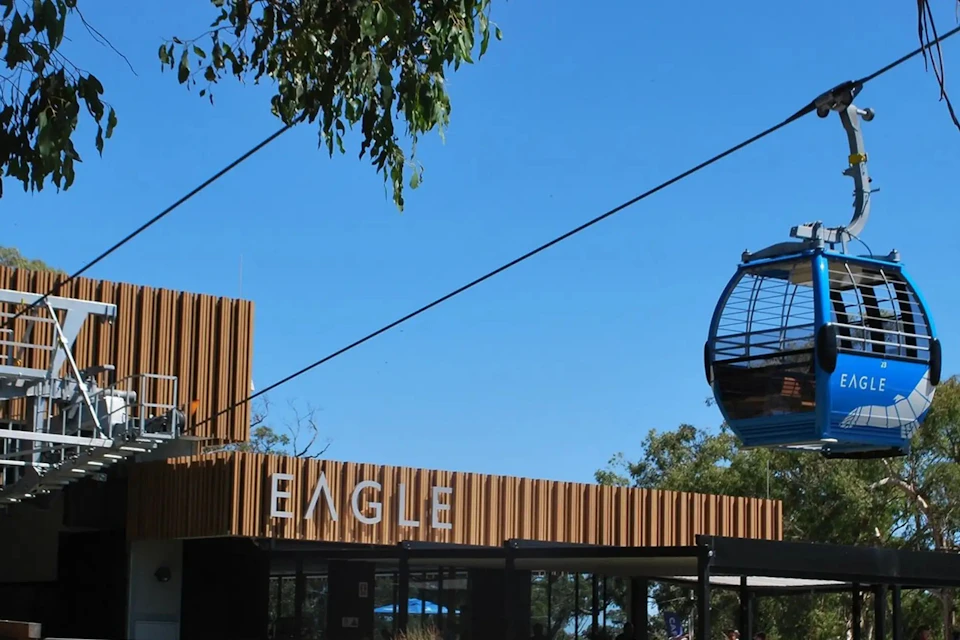 Modern building with "EAGLE" sign and a cable car passing overhead against a clear blue sky. Trees partially visible.