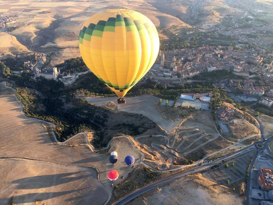 A yellow-hot air balloon floats above a landscape with fields, roads, and buildings. Three smaller balloons are visible below.