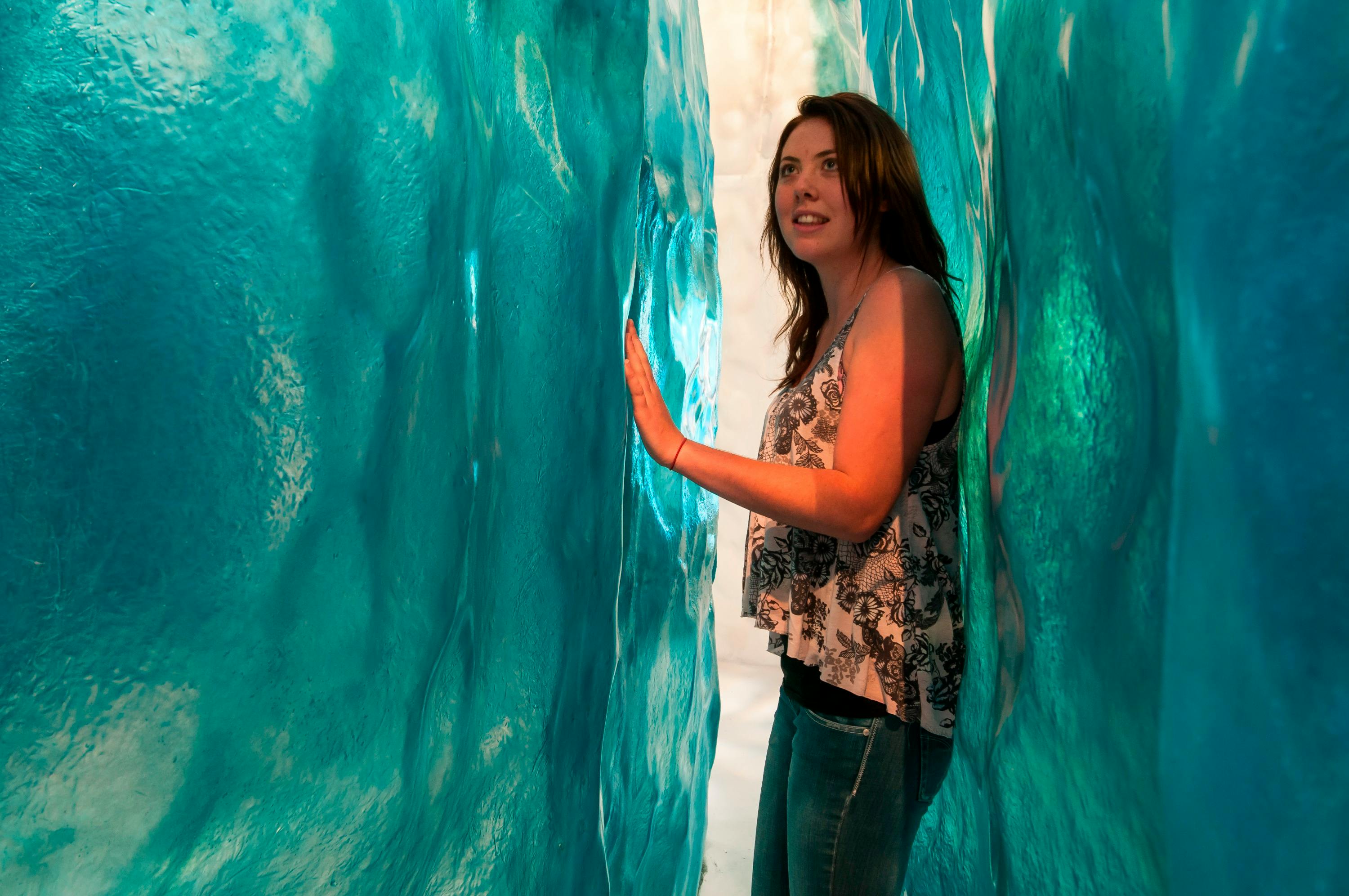 A woman is standing between two large, translucent blue walls, touching one with her hand, and looking ahead.